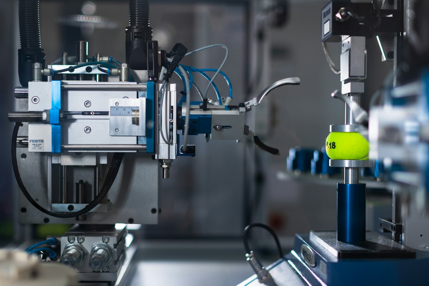 Robotic arm holding a yellow tennis ball near industrial machinery.